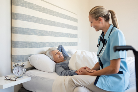 Friendly nurse with senior woman at home. Smiling nurse talking with her patient while getting her out of bed. Disabled woman getting regular health checkup done at home with doctor.の写真素材