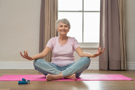 Senior smiling woman doing yoga in her living room. Elderly relaxed woman sitting in lotus pose and meditating while practicing yoga at home. Old grandmother sitting on yoga mat smiling.の写真素材