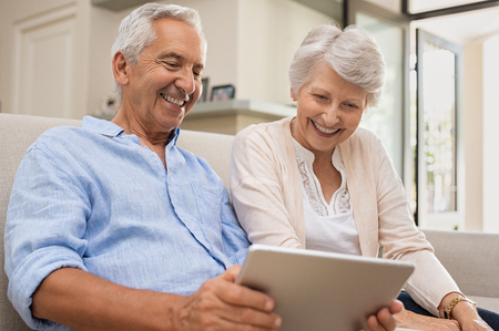 Senior couple using digital tablet sitting on couch at home. Happy old man surfing the net with his wife. Retired man and smiling elderly woman planning on summer vacation using tablet.の写真素材