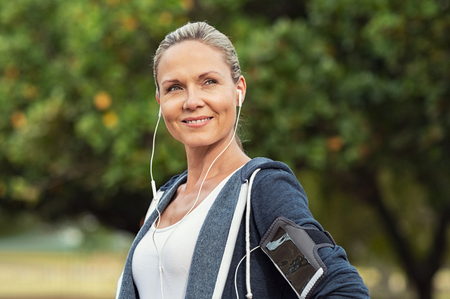 Portrait of a smiling healthy woman with earphones standing in park after running. Proud mature woman looking away after work out. Portrait of satisfied mid woman after fitness exercises.の写真素材
