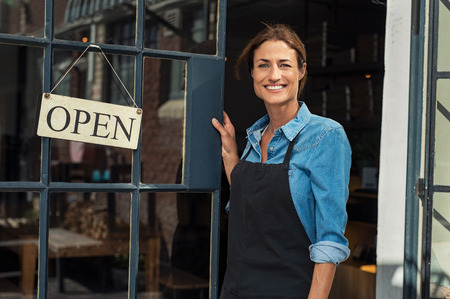 Portrait of a happy waitress standing at restaurant entrance. Portrait of mature business woman in black apron ready to attend new customers in her just opened coffee shop. Happy beautiful woman owner showing open sign in her small business shop.の写真素材