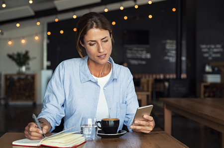 Mature woman sitting in cafe at table and typing a message on smartphone. Middle aged blogger making notes using organizing application on phone. Businesswoman reading information from smartphone while working remotely in cafe interior.の写真素材