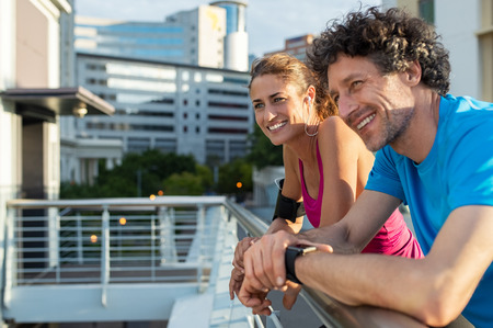 Couple of runners resting while training. Mature woman and man having a rest and smiling while looking away in city. Cheerful healthy couple resting and looking away on railing fence after morning exercise routine.の写真素材