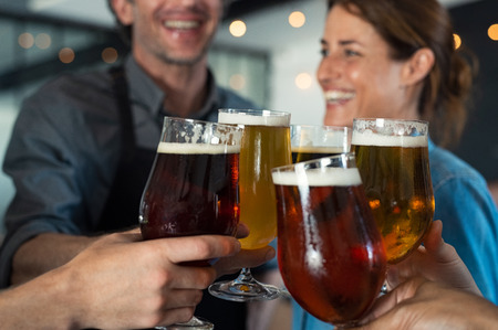 Happy friends clinking glasses of tap beer. Closeup hands of mature men and women toasting with draught beer. Hands holding beer for raising a toast to celebrate. People cheering to success with pints of beer.の写真素材