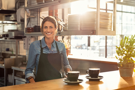 Smiling waitress wearing black apron standing behind counter in cafeteria and looking at camera. Mature woman serving two cups of hot coffee to customer in cafeteria. Small business and entrepreneur concept.の写真素材