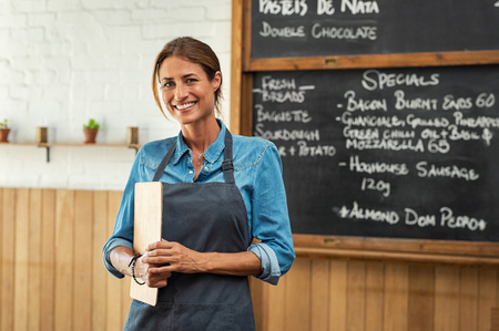 Portrait of smiling mid owner holding clipboard in hand while looking at camera. Beautiful waitress at cafeteria wearing black apron welcoming customers. Mature woman working at small business shop.の写真素材