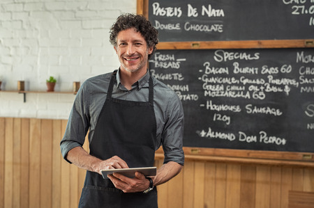 Mature waiter wearing black apron and standing in front of the blackboard with the menu of the day. Portrait of smiling man holding digital tablet and looking at camera. Happy small business owner working in cafeteria with digital tablet.の写真素材