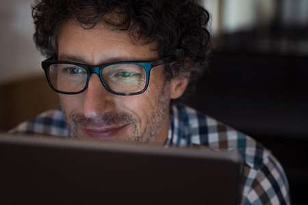 Mature man working on computer at night in dark office. Smiling business man wearing eyeglasses sitting at night working on laptop. Businessman wearing spectacles using digital tablet late at night.の写真素材