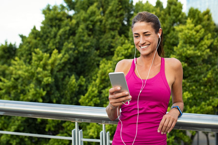 Beautiful mature runner chatting on mobile phone while relaxing after fitness training outside. Smiling woman looking at smartphone screen while listening to music on earphones. Relaxed latin woman resting after jogging in the city.の写真素材