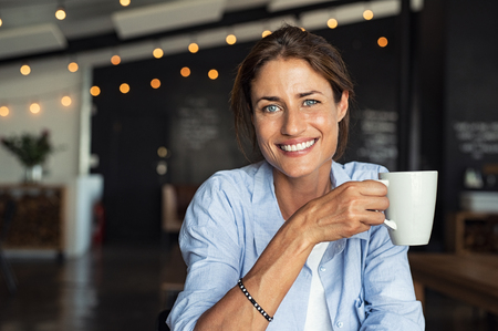Portrait of beautiful mature woman drinking a cup of tea in cafeteria. Happy woman in a cafe drinking coffee and looking at camera. Portrait of smiling lady relaxing in a coffee shop.の写真素材