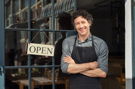 Portrait of small business owner smiling and standing with crossed arms outside the cafe. Portrait of handsome smiling waiter standing in entrance of coffee shop. Successful mature man with arms crossed smiling at camera.の写真素材