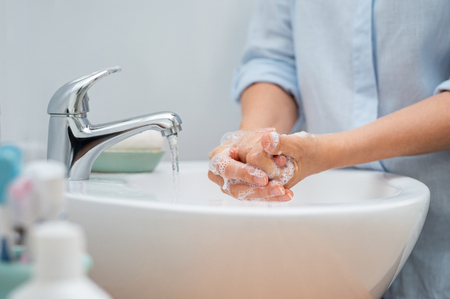 Closeup of woman applying soap while washing hands in basin with open tap. Mature woman washing hands for cleanliness purpose. Lady rubbing hands filled with soap.の写真素材