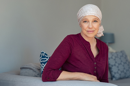 Mature woman with cancer in pink headscarf smiling sitting on couch at home.の写真素材