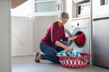 Happy senior woman loading dirty clothes in washing machine. Smiling mature woman sitting on floor putting clothed in washing machine from laundry basket. Housework.