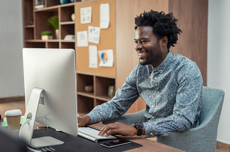 Portrait of african american businessman using computer at desk. Handsome employee enjoying work in modern office. Smiling creative business man feeling happy about positive report.の写真素材