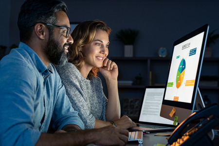 Happy business people working together in office at night. Startup business partners working late together. Mature man and casual woman working with computer late at night in their office.の写真素材