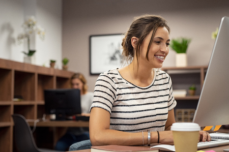 Portrait of smiling young woman working at computer screen. Smiling multiethnic businesswoman working on desktop computer in a modern office. Successful latin girl work in coworking space.の写真素材