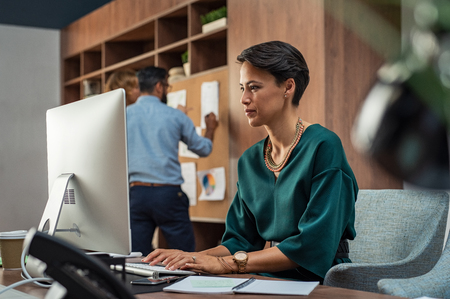 Beautiful businesswoman working on desktop computer in her workstation. Young fashion business woman sitting at desk and using pc with her colleagues in background. Beautiful creative woman sitting on workplace.の写真素材