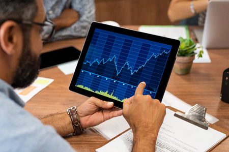 Mature latin businessman reading business graphs and charts on digital tablet. Rear view of business man holding tablet computer with stock analytics and abstract graphs. Closeup of multiethnic casual man hands holding digital tablet with stock market chart.の写真素材