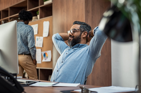 Happy middle eastern businessman relaxing in his office while looking computer monitor. Smiling mature business man with beard and eyeglasses sitting with hands behind head at office desk. Relaxed satisfied casual man taking a break at workplace.の写真素材