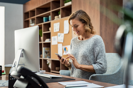 Beautiful smiling woman reading text message on mobile phone during work. Mature businesswoman checking new messages on smartphone. Casual businesswoman checking important mail while working on computer in office.の写真素材