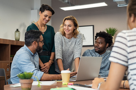 Multiethnic business people talking and smiling during meeting in office. Mature middle eastern man explaining proposal and planning with coworkers. Happy businessman showing project and marketing strategy on laptop to his colleagues.の写真素材