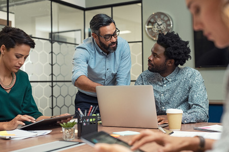 Latin mature manager discussing with his colleague online data in front of laptop. Serious businessman wearing eyeglasses training employees in job. Creative multiethnic team working together with boss in a meeting room.の写真素材