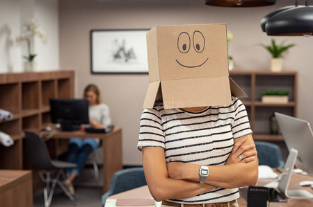 Confident businesswoman wearing positive carton box on head. Young casual woman wearing cardboard box with a happy face standing in office with folded arms. Girl with positive expression drawn on cardboard covering her face.の写真素材
