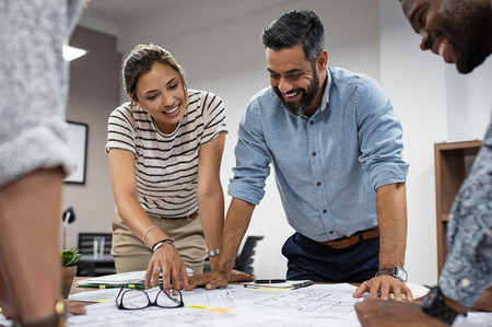 Team of multiethnic architects working on construction plans in meeting room. Cheerful engineers discussing on project in office. Group of mature businessman and young woman standing around table working on blueprint.の写真素材