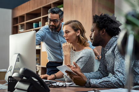 Focused businesspeople discussing project while looking at computer screen. Mature manager working with his creative team in a modern office. Multiethnic casual teamwork at work.の写真素材