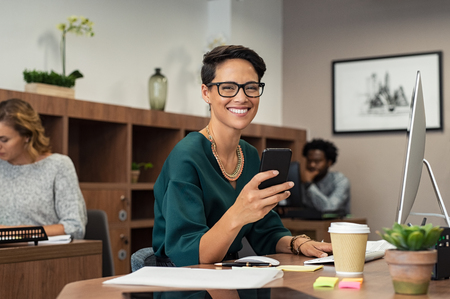 Happy fashionable girl using smartphone while working on desktop computer. Cheerful young business woman sitting in coworking space typing on smart phone and looking at camera. Portrait of  freelancer with eyeglasses working in office.の写真素材