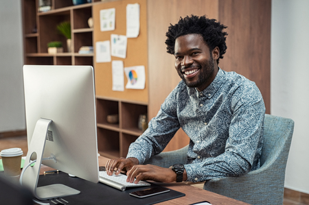 Successful african american businessman typing on desktop computer at modern office. Happy man using computer for work and looking at camera. Portrait of creative business man feeling successful after receiving approval mail from client.の写真素材