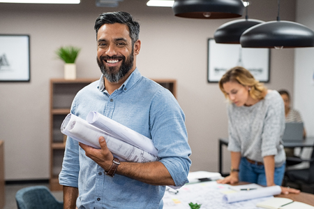 Portrait of multiethnic architect with blueprints in creative office. Mature middle eastern contractor holding roll of architectural projects while looking at camera. Happy latin man in casual standing in meeting room with engineers working in background.の写真素材