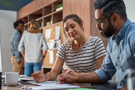 Mature businessman and young businesswoman discussing work and writing notes in office. Creative business partners working together sitting at desk. Middle eastern manager wearing spectacles and helping woman with company details.の写真素材