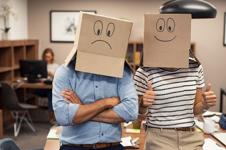Business man and businesswoman covering face with cardboard box showing happy and sad face. Businessman with folded arms wearing sad face near his colleague with positive expression. Young woman with thumbs up gesture sign at office.の写真素材