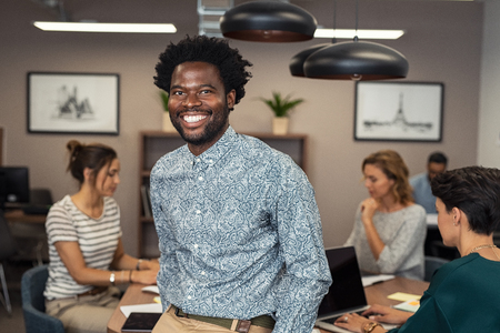 Portrait of happy african businessman looking at camera and sitting at desk. Mature business man in office with colleagues working in background. Successful african american man in casual standing with his colleagues at office.の写真素材