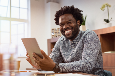 Happy casual african man using digital tablet at office. Portrait of smiling black businessman sitting on chair using tablet pc. African american casual business man looking at camera.の写真素材