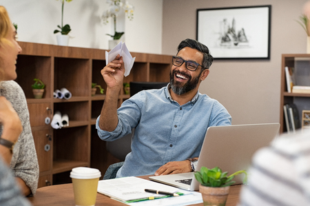 Cheerful mature business man playing with paper plane in office. Team of designers having fun while middle eastern man throwing plane on colleagues. Portrait of happy smiling businessman with eyeglasses and beard enjoying with colleagues in a meeting room.の写真素材