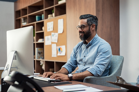 Smiling creative business man typing on desktop computer in office. Mature middle eastern businessman working at modern office space. Happy hispanic man wearing casual and eyeglasses working on his computer.の写真素材