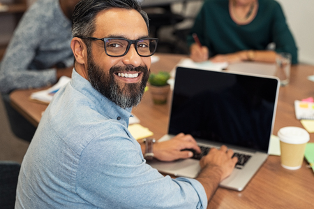 Mature middle eastern businessman using laptop at desk in busy office. Confident business man in a creative agency with team in background. Happy smiling casual man looking at camera while sitting at table and working on laptop computer.の写真素材
