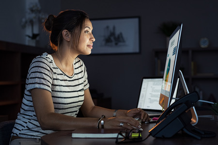 Young businesswoman in casual clothing working on desktop computer making business plan. Beautiful tired business woman working on assignment at night. Latin girl using computer late at night in office.の写真素材