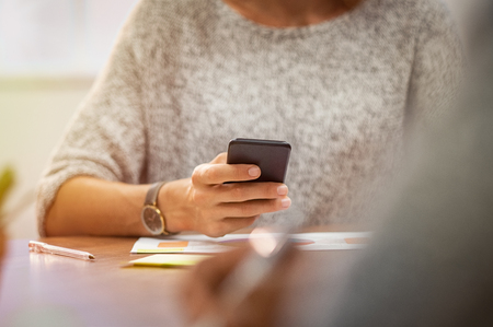 Closeup of woman hands holding smartphone during meeting. Woman reading message in mobile phone while sitting in office. Casual businesswoman hand using smart phone and typing on it.の写真素材