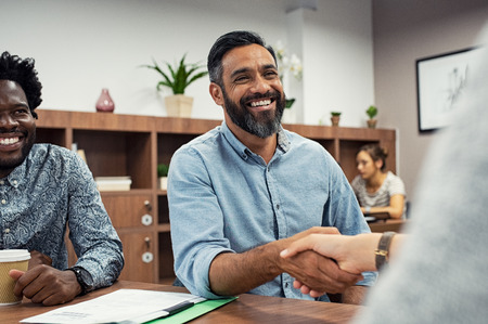 Two business people shaking hands while sitting in meeting room. Middle eastern businessman shake hands to businesswoman. Portrait of happy smiling latin man signing off deal with an handshake.の写真素材