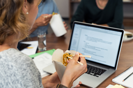 Business woman eating instant noodles with chopsticks while working at office. Casual businesswoman reading document on laptop while eating take away food. Executive working during lunch break with colleagues in background.の写真素材