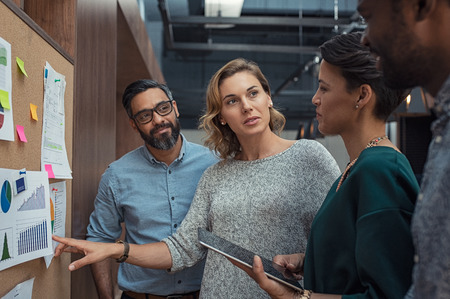 Multiethnic business people discussing charts and graphs showing the results of their successful teamwork. Mature business woman showing presentation to casual colleagues in modern office during night meeting. Group of creatives analyzing market trends.の写真素材