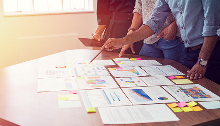 Businessman and business woman discussing on stock market charts in office. Closeup of business hands in meeting room discussing on graphs. Multiethnic man pointing pencil at document in conference room with his colleagues around the table.の写真素材