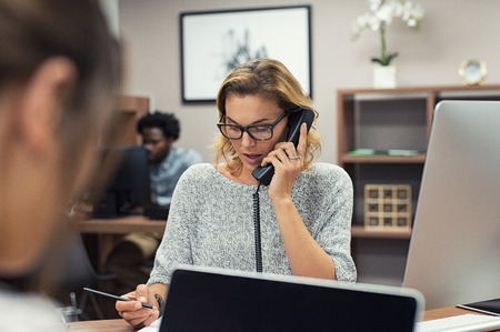 Mature businesswoman talking on phone in office. Casual business woman talking on landline phone in a creative agency. Beautiful blonde receptionist with eyeglasses taking client details over phone.の写真素材