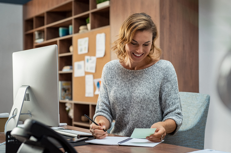 Smiling busy employee in casual reading notes and writing on notebook. Happy mature business woman reading letter and smiling at office. Creative businesswoman writing in book with pencil while taking notes for meeting.の写真素材