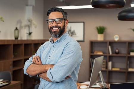 Portrait of happy mature businessman wearing spectacles and looking at camera. Multiethnic satisfied man with beard and eyeglasses feeling confident at office. Successful middle eastern business man smiling in a creative office.の写真素材