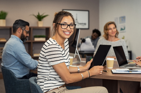 Beautiful latin businesswoman looking at camera and smiling while working in office. Cheerful young woman wearing spectacles and holding digital tablet. Creative girl working with colleagues at agency.の写真素材